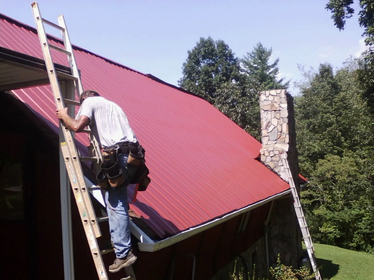 Roofer on a ladder beside a red metal roof and stone chimney — Soffit & Fascia Repair by Roofing Brothers in Ruffin